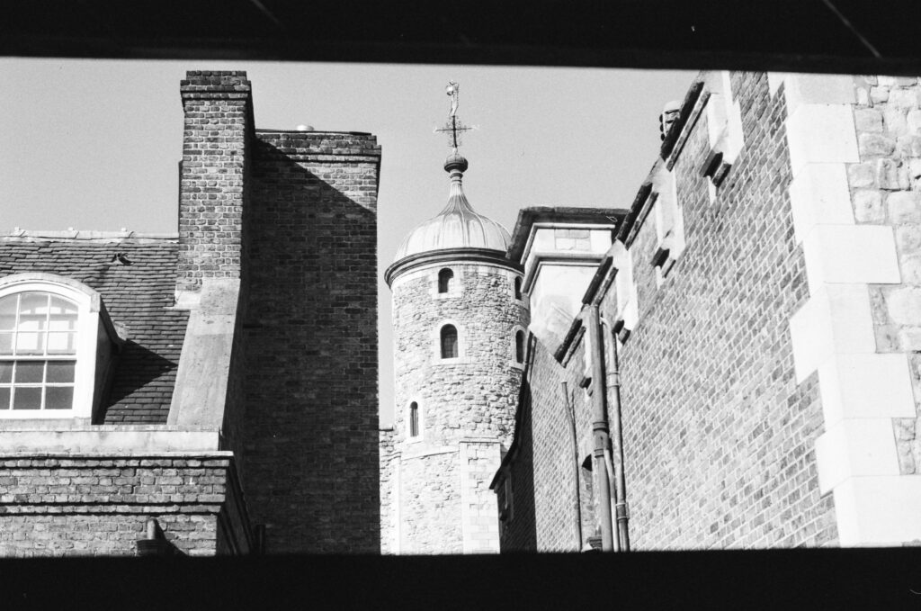 Black and white photo of old brick buildings with a round stone tower topped by a weather vane, framed by dark edges at the top and bottom.