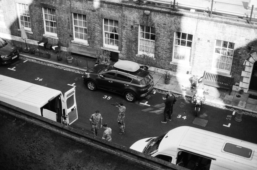 Black and white image of a street with parked cars and vans. Four people in uniform stand near the vehicles, with a brick building and several windows in the background. The ground is marked with numbered parking spaces.