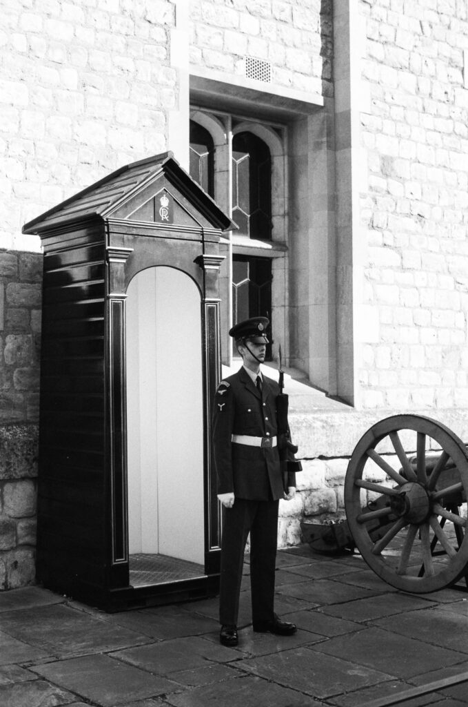 A uniformed guard stands at attention beside a small wooden sentry box and an old cannon, in front of a stone building.