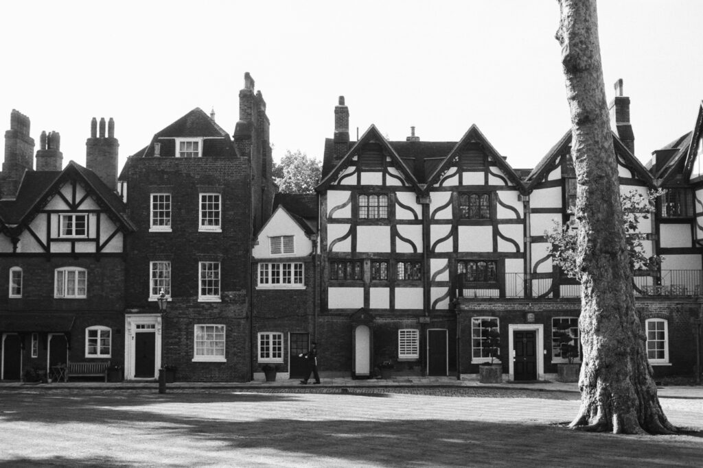 Black and white photo of old timber-framed houses with gabled roofs and tall chimneys. A large tree stands in the foreground, and a person walks on the lawn in front of the buildings.