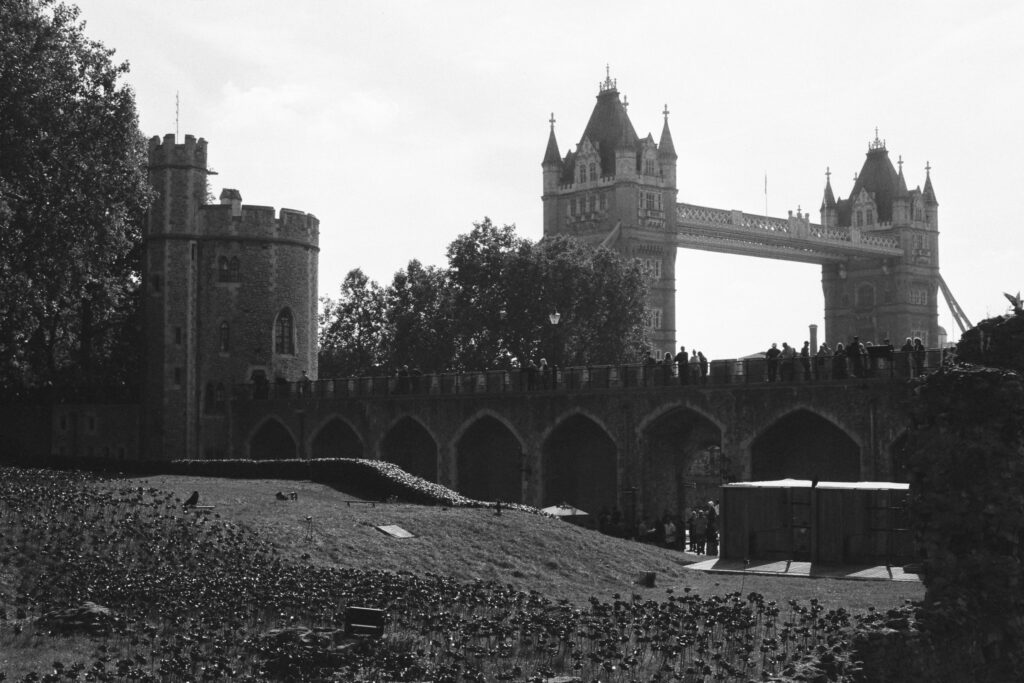 Black and white photo of Tower Bridge in London, with a historic stone building and poppy display in the foreground. Silhouetted people are visible on the bridge. Trees and cloudy sky in the background.