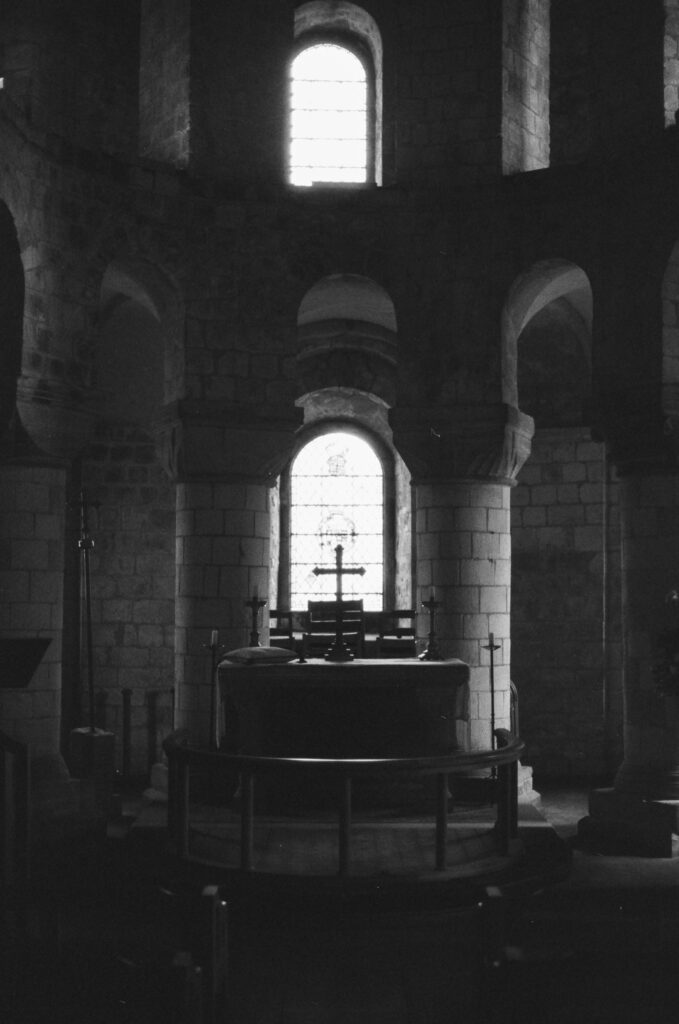 A black-and-white photo of a church altar with a cross, tall arched windows behind it letting in light, and stone walls and pillars surrounding the altar.