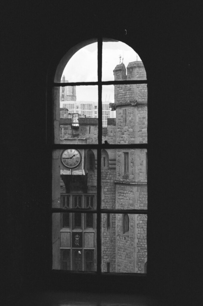 A black-and-white photo of a stone building with a clock, seen through a tall arched window with four panes. Modern buildings and a cloudy sky appear faintly in the background.