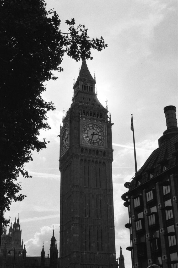 Black and white photo of Big Ben clock tower in London, partially framed by tree branches and nearby buildings, with the sky in the background.
