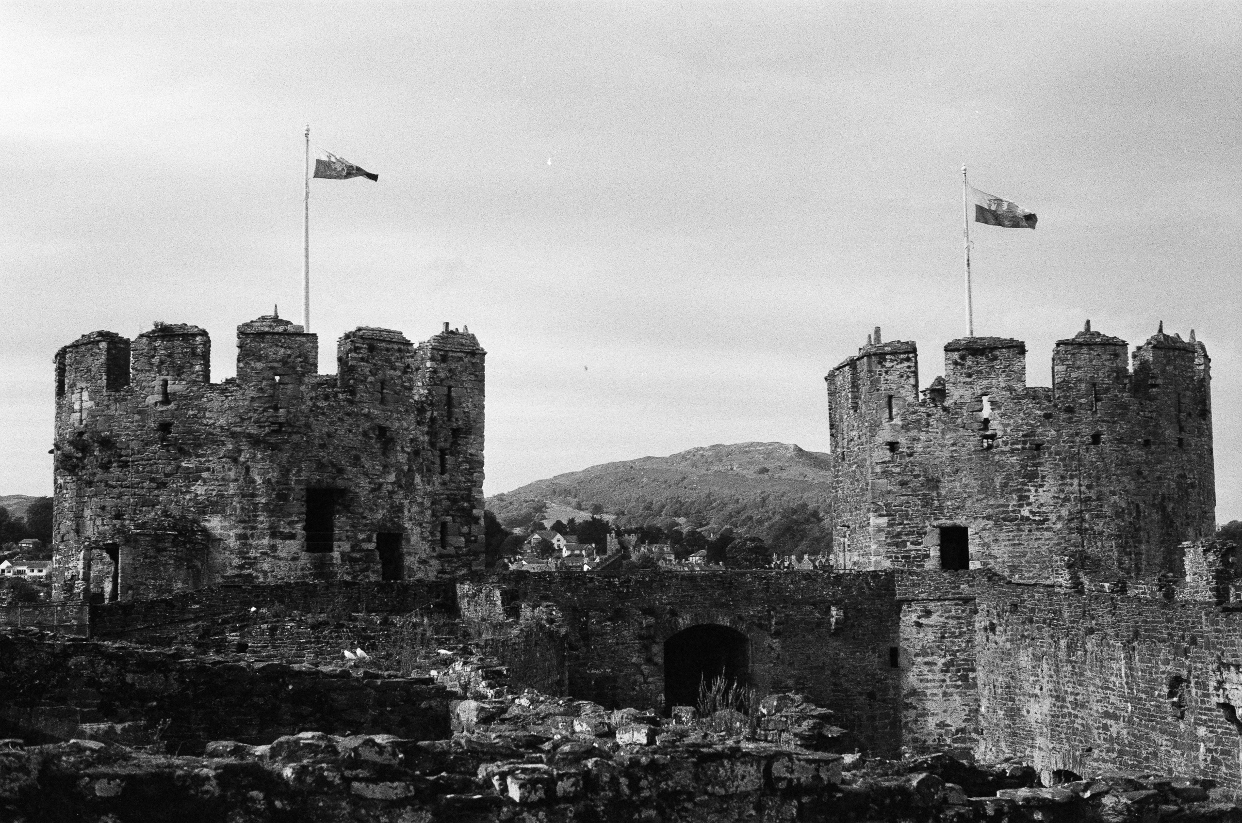 Two stone towers of a castle ruin, each topped with a flag, overlooking a town and hills.