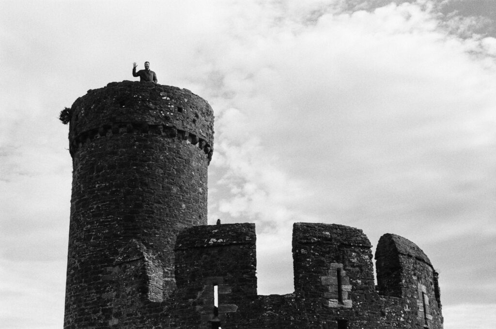A person stands on top of a stone tower waving, against a cloudy sky.
