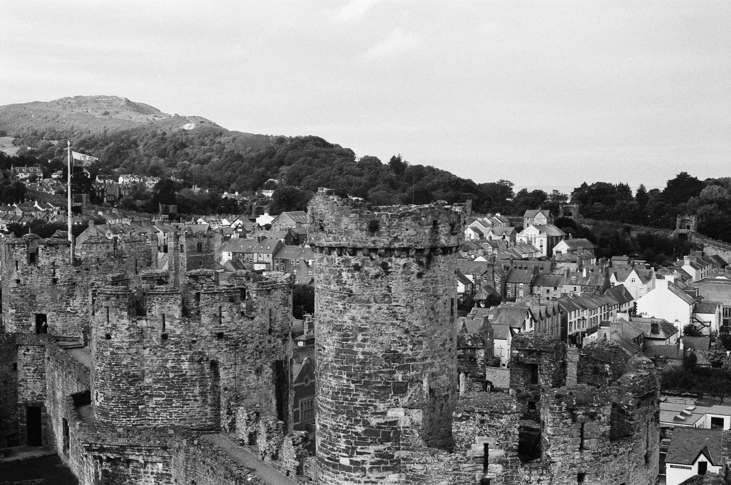 A black and white high-angle view of stone castle ruins overlooking a town nestled at the foot of a forested hill.
