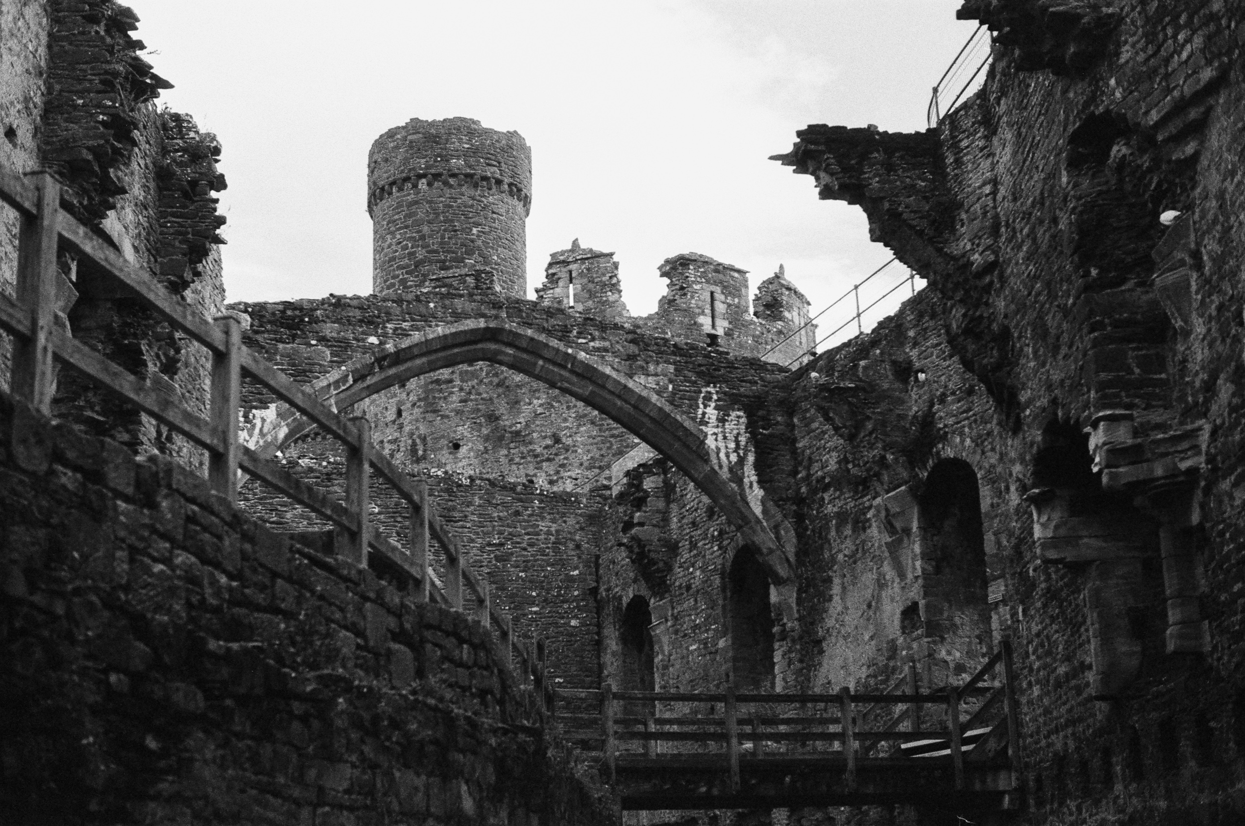 Stone ruins of a castle featuring a central tower, an arched stone gateway, and wooden walkways.