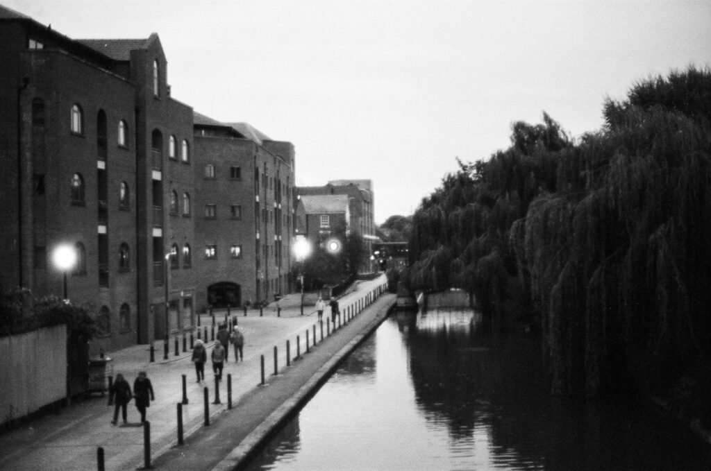 People walking along a canal path lined with multi-story brick buildings and weeping willow trees.