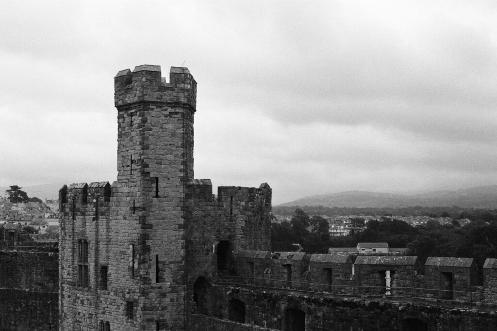 Stone castle tower and crenelated walls overlooking a town under a cloudy sky.