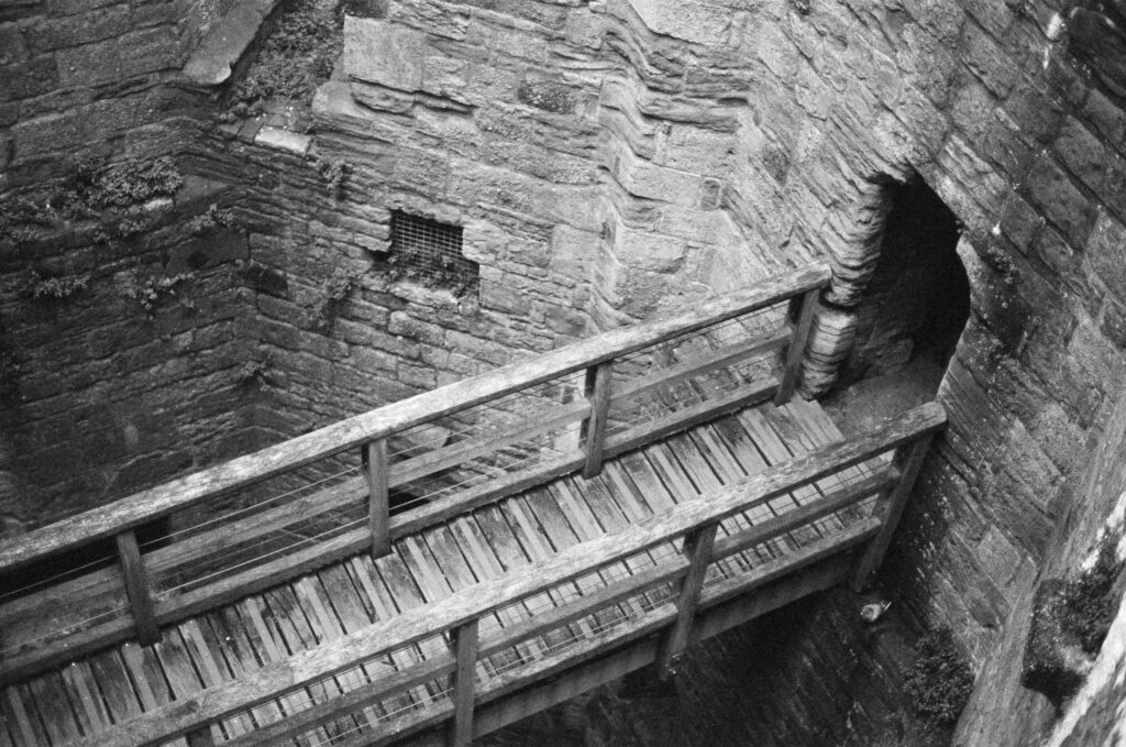 A wooden bridge with railings crosses a stone chasm toward a dark arched doorway in a stone wall.