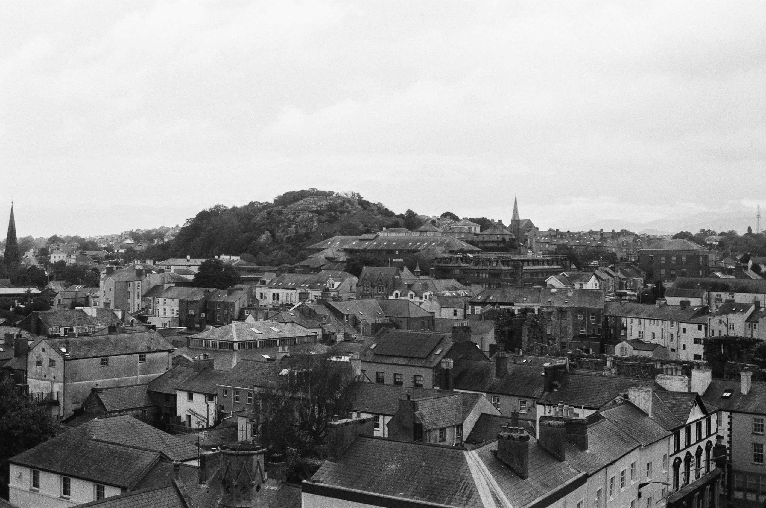 A black and white high-angle view of a dense town with traditional buildings, tiled roofs, and a wooded hill in the distance.