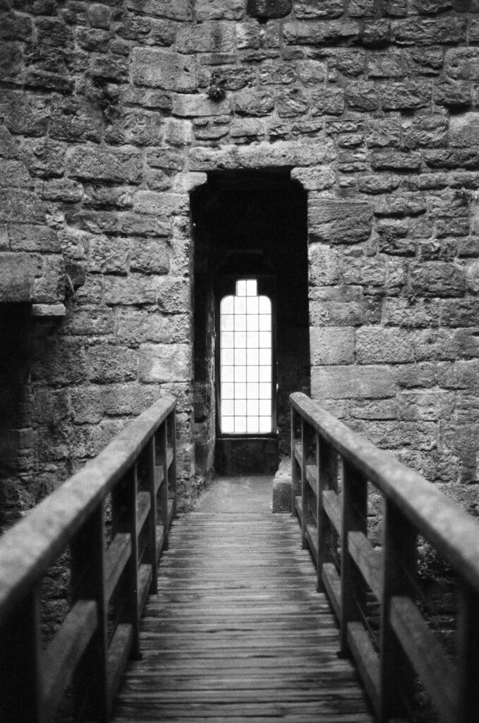 A wooden footbridge with railings leads toward a tall, narrow window set in a thick stone wall.